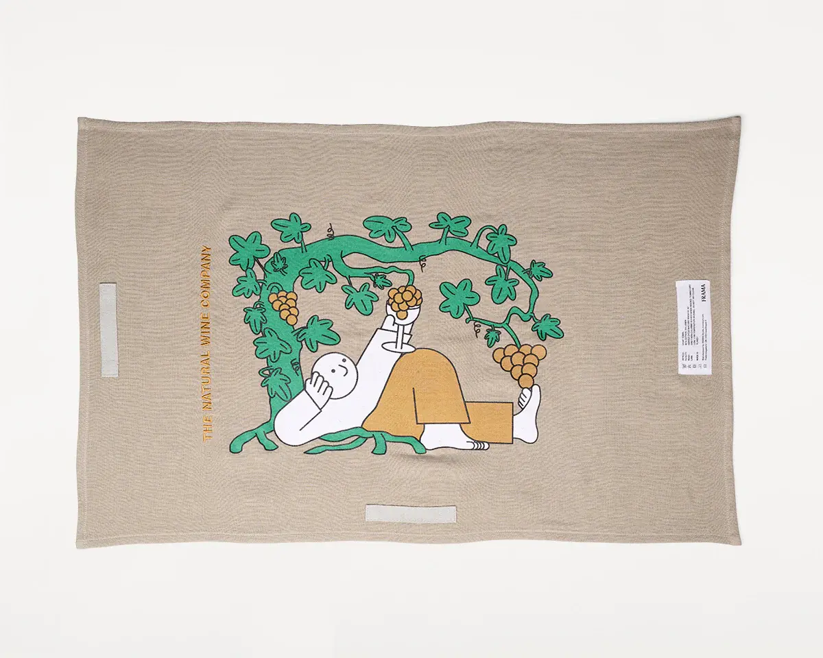 Tea towel from a collaboration between Jhon Boy, Frama, and The Natural Wine Company (TNWC). The design features a playful illustration of a relaxed figure reclining under a lush grapevine, holding a bunch of grapes. The text ‘The Natural Wine Company’ is printed on the side, combining Jhon Boy’s minimalistic style with the refined aesthetics of Frama and TNWC.