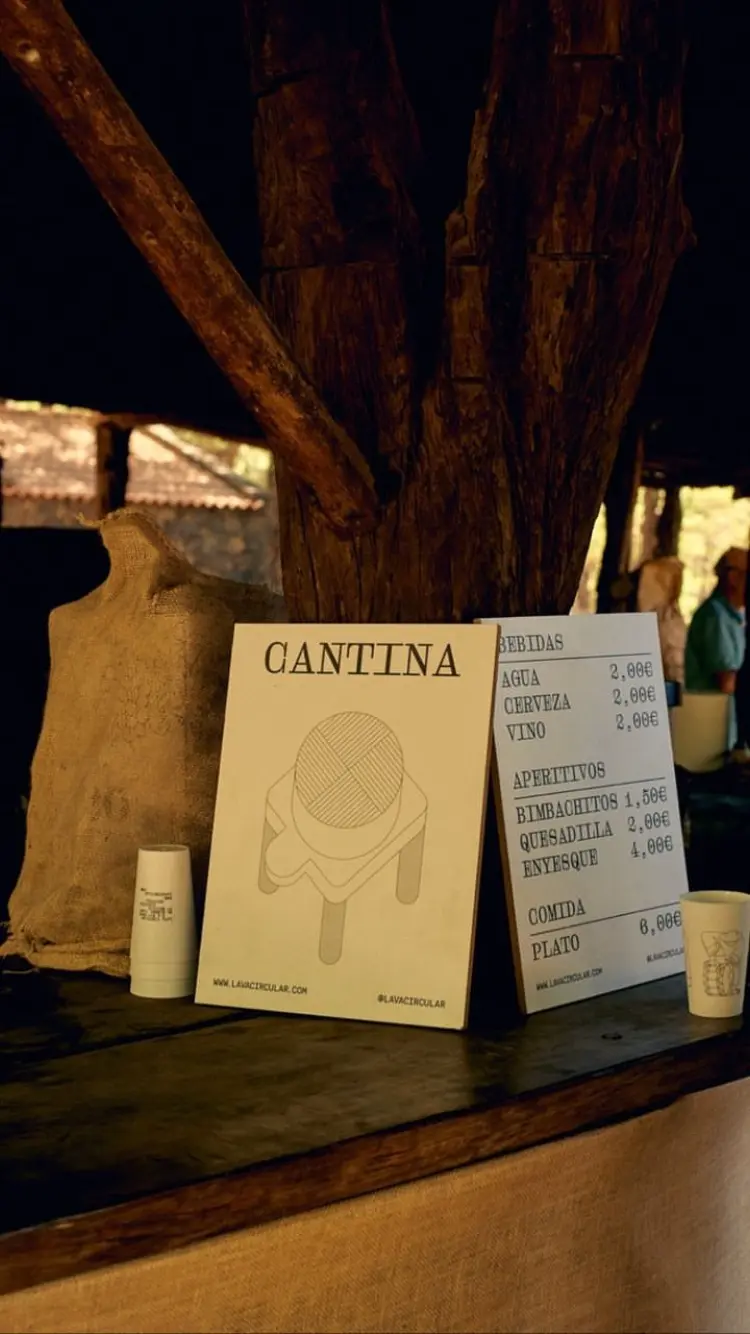 A rustic cantina setup with a Lava Circular poster featuring an illustrated figure playing a traditional drum, accompanied by a menu listing food and beverage options. Cups with matching illustrations are also present, tying the visual identity together. The natural wooden textures complement the cultural theme of the event, reinforcing the brand identity created by Jhon Boy. Photograph by Adrian Rios.