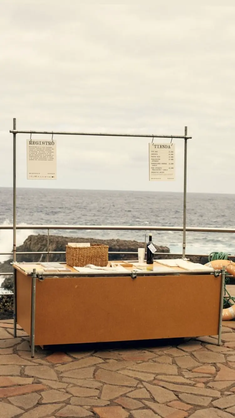 A registration desk by the ocean for the Lava Circular event. Two posters are displayed above the desk: one reads “Registro” with event details, and the other lists items for sale under “Tienda.” The table is simple, featuring materials like woven baskets and brochures, blending with the serene ocean background. This image highlights the minimalistic setup of the Lava Circular event, part of the brand identity designed by Jhon Boy. Photograph by Adrian Rios.