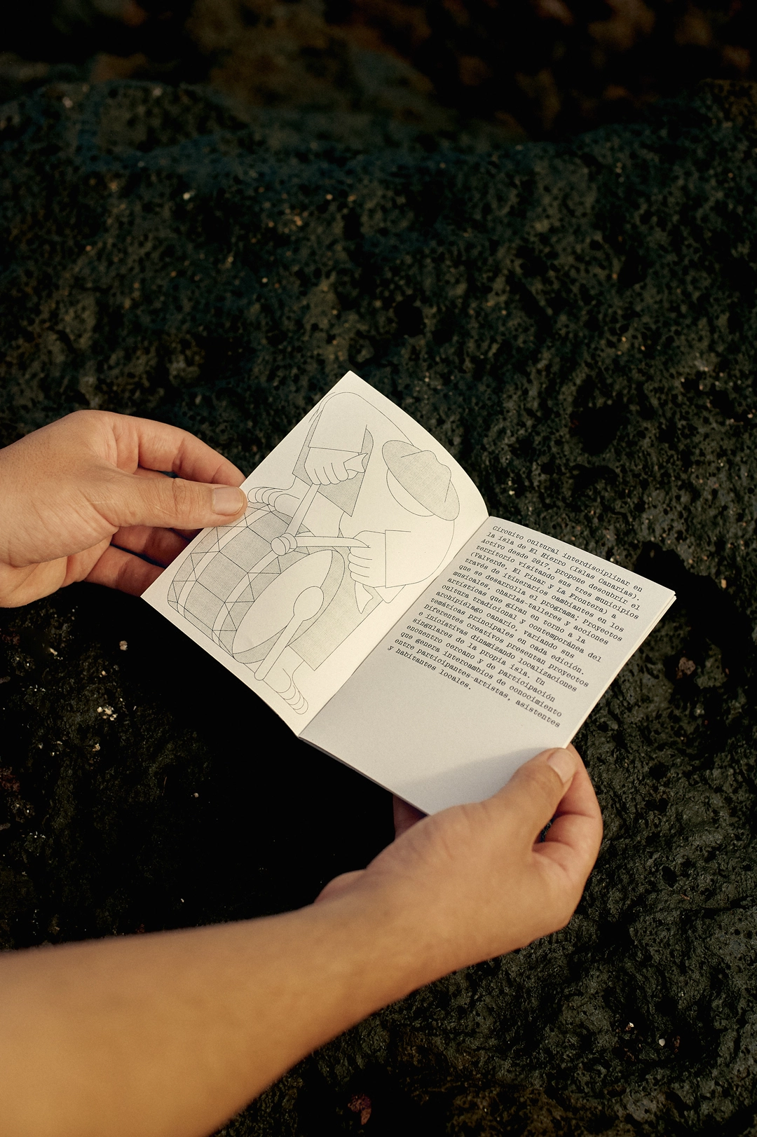 A person flipping through a booklet with a Jhon Boy illustration of a figure playing a drum. The text in Spanish details the Lava Circular event’s cultural significance. The booklet is placed on volcanic rock, emphasizing the connection to El Hierro’s natural environment and the event’s essence. Photograph by Adrian Rios.